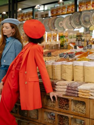 Two stylish women in a market showcasing fashion amidst colorful spices and grains.