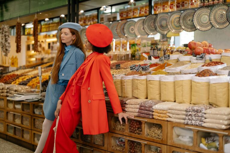 Two stylish women in a market showcasing fashion amidst colorful spices and grains.